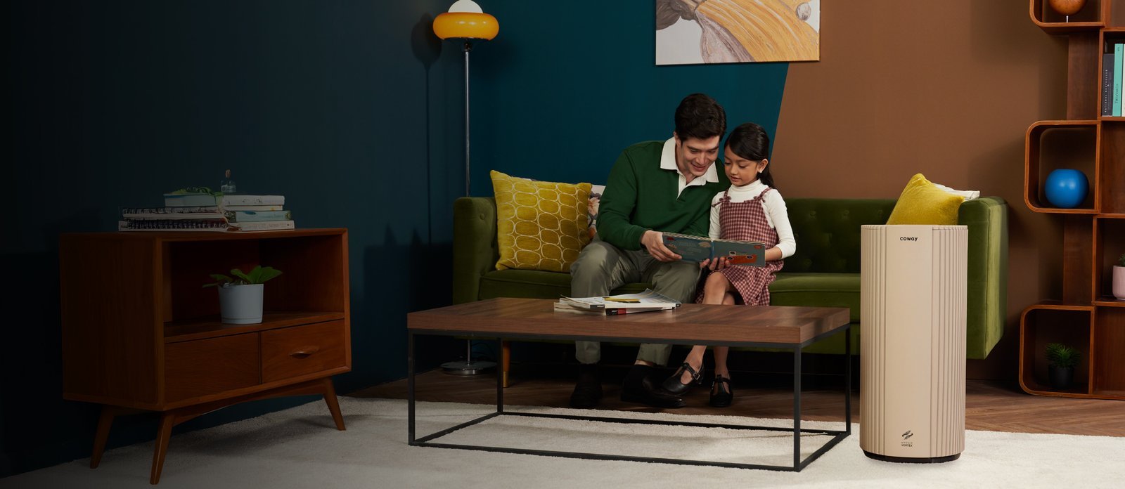 A father and daughter reading together on a green sofa with the Coway Suite air purifier beside them