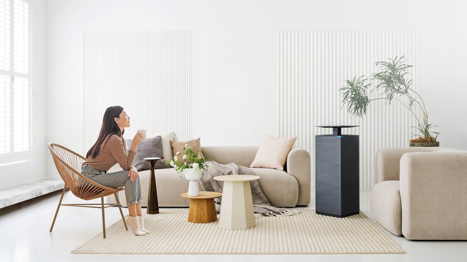 A woman relaxing with a warm drink in a bright living room beside the Coway Noble air purifier